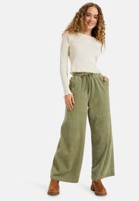 Vrouw met krullend haar, gekleed in een crèmekleurig langemouwen top en losse olijfgroene corduroybroek met een paar bruine enkellaarsjes, lachend.