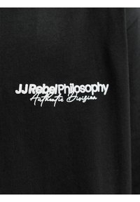 Μαύρο ύφασμα με λευκό κείμενο «JJ Rebel Philosophy Authentic Division» κεντημένο πάνω του.