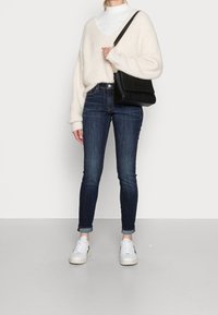 Persona vestida con un suéter de cuello alto blanco y un suéter de pico esponjoso, jeans oscuros de mezclilla remangados, zapatillas blancas y llevando un bolso negro de hombro.