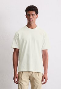 Helles gelbes Baumwoll-T-Shirt mit kurzen Ärmeln, Rundhalsausschnitt und lässiger Passform, kombiniert mit beigen Hosen vor einem neutralen Hintergrund.