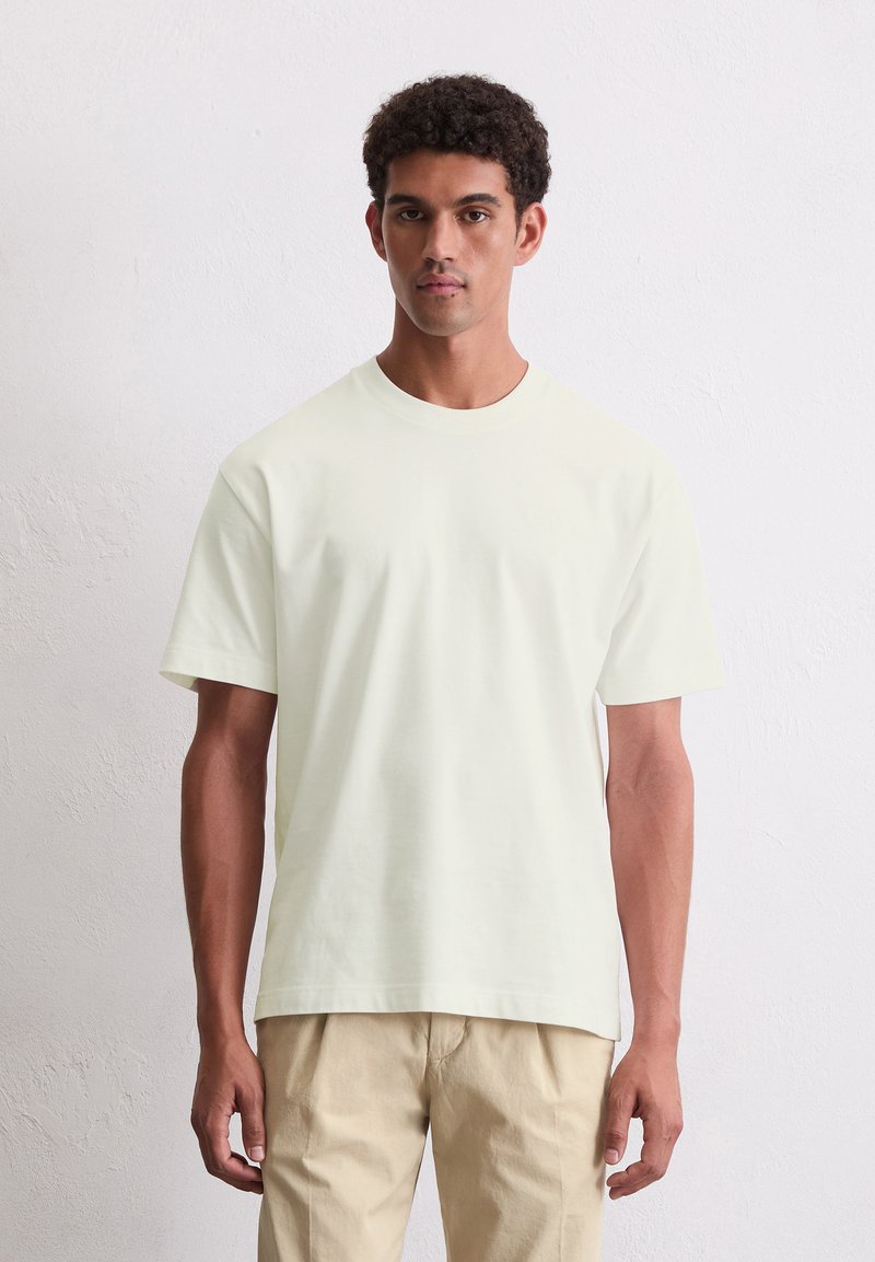 Helles gelbes Baumwoll-T-Shirt mit kurzen Ärmeln, Rundhalsausschnitt und lässiger Passform, kombiniert mit beigen Hosen vor einem neutralen Hintergrund.