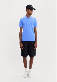 Lys blå polo-skjorte med korte ermer, med logo, kombinert med svarte cargo-shorts og hvite sneakers. Minimalistisk design.