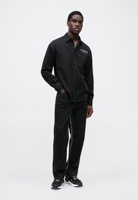 Tricou negru cu mânecă lungă, cu închidere cu nasturi și un buzunar pe piept; asortat cu pantaloni negri cu dungi albe pe laterale; purtat cu adidași negri.