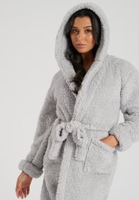Robe en peluche gris avec une capuche, dotée d'une taille à nouer et de deux poches latérales. Sa texture douce et sa surface duveteuse lui confèrent une apparence confortable.