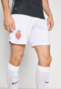 Un short de sport blanc présente un logo AS Monaco doré avec une couronne. Associé à un maillot noir et des chaussettes blanches avec un swoosh Nike noir.