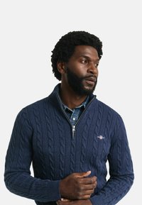 Navy-blauer Kabelstrick-Pullover mit halbseitigem Reißverschlusskragen, gerippten Bündchen und dezentem Logo-Detail auf der Brust, über einem Jeanshemd getragen.