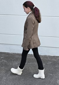 Blazer à carreaux marron, jean noir slim et bottes en caoutchouc blanches. Le blazer présente un col cranté et une coupe ajustée.