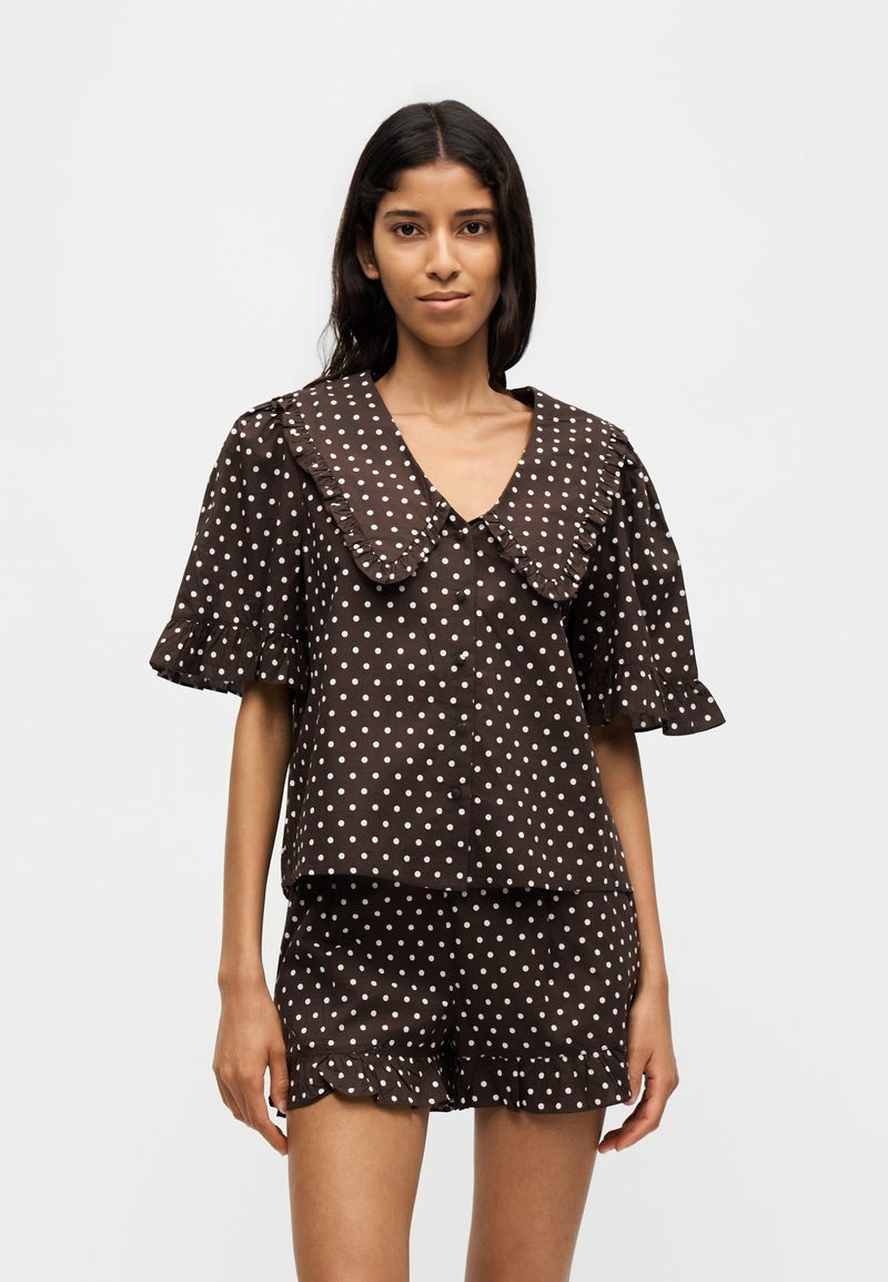 Femme portant un chemisier marron à pois à manches courtes avec un grand col et un short assorti aux ourlets volantés, debout devant un fond uni.