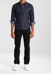 Ciemnoniebieska koszula dżinsowa zapinana na guziki, z dwoma kieszeniami na piersi, rękawy podwinięte; w zestawie z czarnymi spodniami i białymi trampkami.