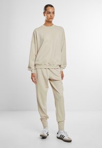 Beige genser og matchende joggebukser laget av myk stoff. Løs passform med ribbestrikkede mansjetter og midjebånd. Hvit sneakers med svarte striper.