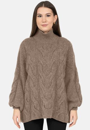 Woman wearing oversized brown cable-knit turtleneck sweater with balloon sleeves against plain background.