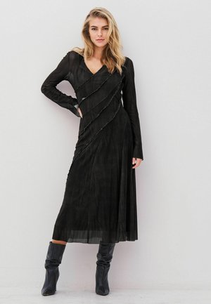 Schwarzes Langarmkleid mit V-Ausschnitt, das wellenförmige Rüschendetails an der Vorderseite aufweist. Kombiniert mit kniehohen schwarzen Stiefeln. Glatte Textur.