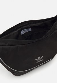Borsa a spalla nera adidas con texture liscia, chiusura con zip e logo. Presenta strisce grigie a contrasto e accenti a scacchi sui lati.