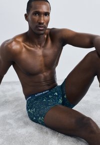 Tamsiai mėlynos boxer tipo apatinės kelnės su žalių ir raudonų gėlių raštu. Yra ženklinta juosta. Modelis sėdi ant šviesios tekstūruotos paviršiaus.