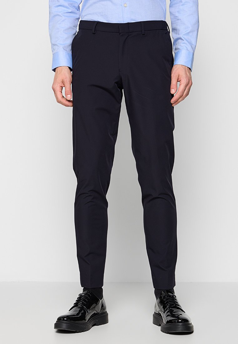 Mann in schlanken schwarzen Anzughosen, hellblauem Langarmhemd, schwarzen Socken und glänzenden schwarzen Dressschuhen, der vor einem weißen Hintergrund steht.