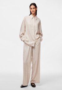 Camicia oversized a righe beige con taschino sul petto, abbinata a pantaloni larghi coordinati. Texture liscia, vestibilità rilassata, stile casual.