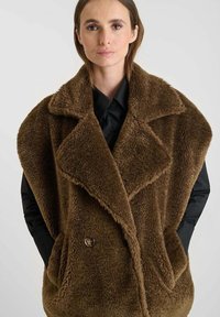 Bruine nep-bont vest met een brede kraag, dubbele knoopsluiting, grote zakken en een losse pasvorm, gedragen over een zwart shirt.