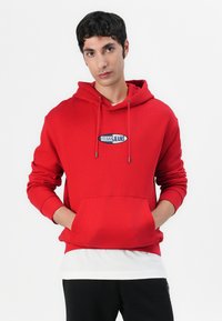 Red hoodie with a front pocket, drawstring hood, and embroidered "GUESS JEANS" logo in an oval. Textured fabric and relaxed fit.