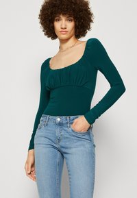 Teal langermet body med firkantet utringing, drapert detalj ved brystet og glatt stoff, sydd sammen med lyseblå jeans.