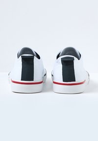 Hvite kanvas sneakers med en svart stoffheltapp, en teksturert hvit gummsåle og et rødt aksent på yttersålen.