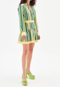 Vestido a rayas en verde, amarillo y blanco, con cuello camisero y falda de volantes. Combinado con zapatos de plataforma gruesa de color verde brillante.