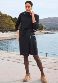 Zwarte lange mouwen top met een hoge kraag en knopdetail, gecombineerd met een zwarte jurkrok van faux leer en tan enkelboots.