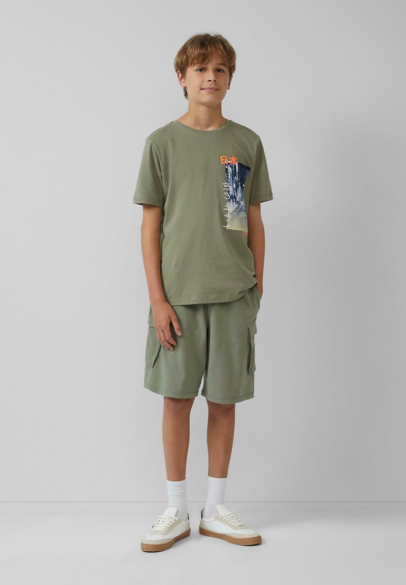 Olijfgroene katoenen t-shirt met grafische print, gecombineerd met bijpassende cargoshorts. Witte sneakers en enkelsokken maken de outfit compleet.