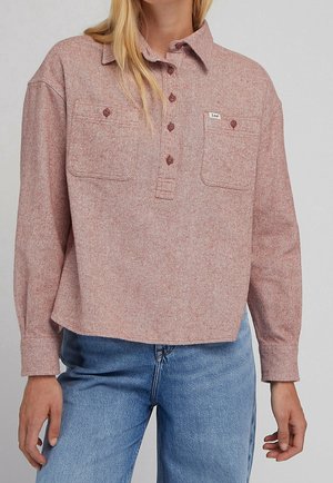 Personne portant une chemise rouge clair à manches longues avec deux poches poitrine et un jean bleu, debout devant un fond uni.