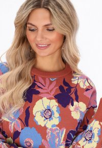 Donna con lunghi capelli biondi mossi che indossa un maglione color ruggine con grandi motivi floreali gialli, blu, rosa e viola, sorridente e con lo sguardo rivolto verso il basso.