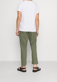 Persoon in een wit shirt met korte mouwen, olijfgroene cropped broek en zwarte open sandalen, staand tegen een effen lichte achtergrond.