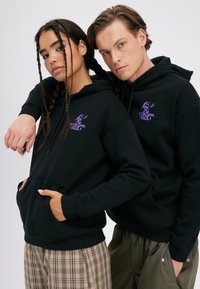 Mladi moški in ženska nosita ujemajoče se črne puloverje s kapuco z vijoličnim napisom "Nikoli ne glej nazaj", stojita skupaj z rokami v žepih na belem ozadju.