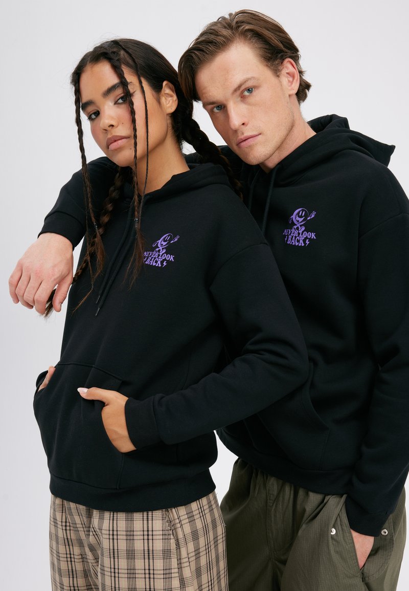 Mladi moški in ženska nosita ujemajoče se črne puloverje s kapuco z vijoličnim napisom "Nikoli ne glej nazaj", stojita skupaj z rokami v žepih na belem ozadju.