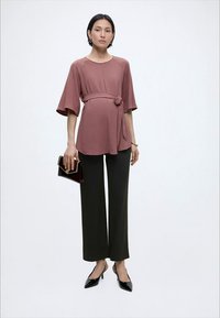 Naine on seljas tolmroosa vööga pluus ja mustad laiad püksid, hoides tumedat käekotti ja kandes musti teravate ninadega kontsakingi.