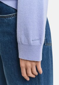 Helle lila gestrickte Pulloverwärmer mit geripptem Bündchen; verfügt über ein kleines, im gleichen Farbton gesticktes Logo. Hand ruht auf Denim-Stoff.