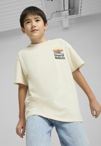 Camiseta color crema de manga corta con un estampado gráfico del texto "PUMA Board Rebels" y un logo, combinada con jeans de color azul claro.