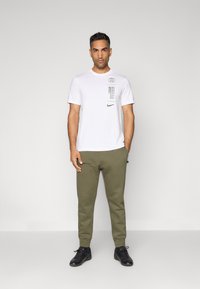 T-shirt branca de mangas curtas com grafismos impressos, combinado com calças de treino verde azeitona que apresentam um logo. Tênis esportivos pretos completam o visual.
