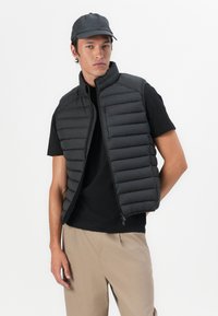 Sort puffer vest med vandret quiltet mønster og høj krave. Lynlås lukning og sidelommer. Båret over en sort t-shirt.