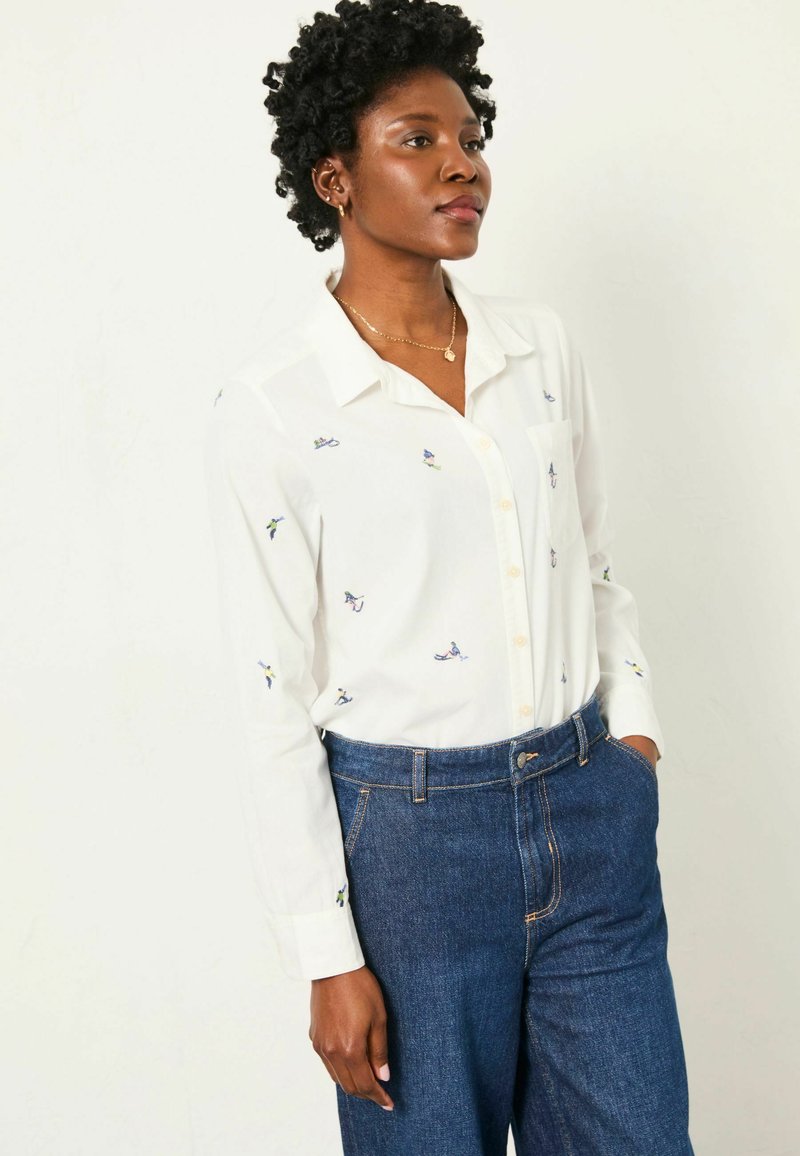 White button-up shirt with colorful embroidered designs. Soft material, long sleeves, and a chest pocket, paired with dark blue denim pants.