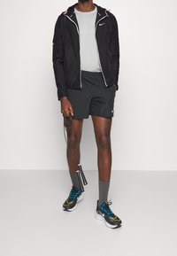 Mężczyzna w czarnej kurtce Nike, czarnych szortach, szarych skarpetach i wielokolorowych butach do biegania, trzyma czarną skakankę w jednej ręce.