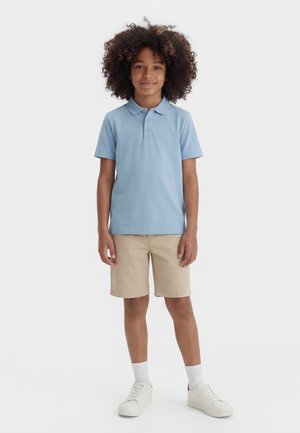 Persona joven con cabello rizado de pie mirando al frente, vestida con una camiseta polo azul claro, shorts beige, calcetines blancos y zapatillas blancas.