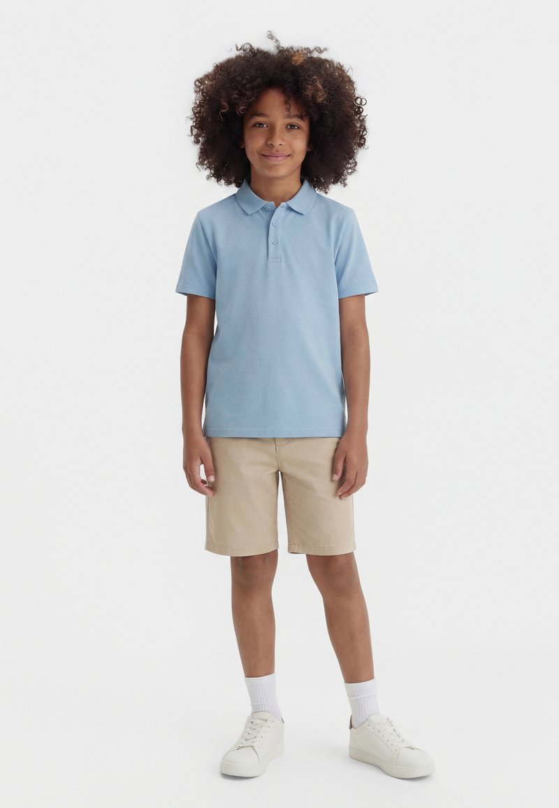 Νεαρό άτομο με κατσαρά μαλλιά στέκεται όρθιο κοιτώντας μπροστά, φοράει ανοιχτό μπλε πόλο, μπεζ σορτς, λευκές κάλτσες και λευκά αθλητικά παπούτσια.