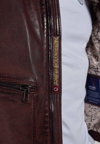 Veste en cuir marron avec fermeture éclair à l'avant, finition texturée, broderies dorées avec "Patrouille de France" et doublure à motifs.