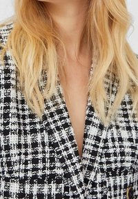 Blont hår som faller över en svartvit rutigt blazer med en djup V-ringning.