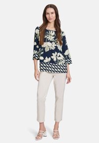 Granatowa bluzka w kwiaty z kremowymi i zielonymi wzorami, szerokim dekoltem w serek i rękawami o długości trzech czwartych; zestawiona z jasnobeżowymi, tapered spodniami.