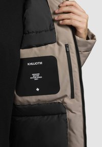 Unutrašnjost puffer jakne u bež i crnoj boji, sa zip džepom i oznakom koja glasi "KHUJOTM GENUINE BRAND ESTABLISHED 2001."