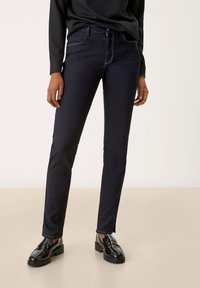 Personne portant un jean slim-fit bleu marine foncé et des mocassins noirs brillants, se tenant sur un sol beige clair devant un fond uni.