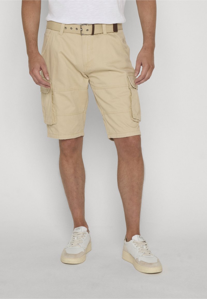 Homme portant un short cargo beige avec des poches latérales, une ceinture marron, des baskets blanches et une chemise blanche devant un fond uni.