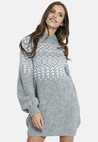 Uśmiechnięta kobieta ubrana w luźną szarą dzianinową sukienkę z białymi geometrycznymi wzorami na piersiach oraz długimi bufkowatymi rękawami.