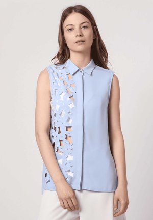 Joven mujer con camiseta sin mangas azul claro con patrón floral recortado en un lado, posando frente a un fondo neutro.
