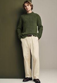 Oliwkowy sweter w prążki, kremowe spodnie, ciemnobrązowe buty, luźny krój, minimalistyczny design, teksturowany materiał, stojący na neutralnym tle.